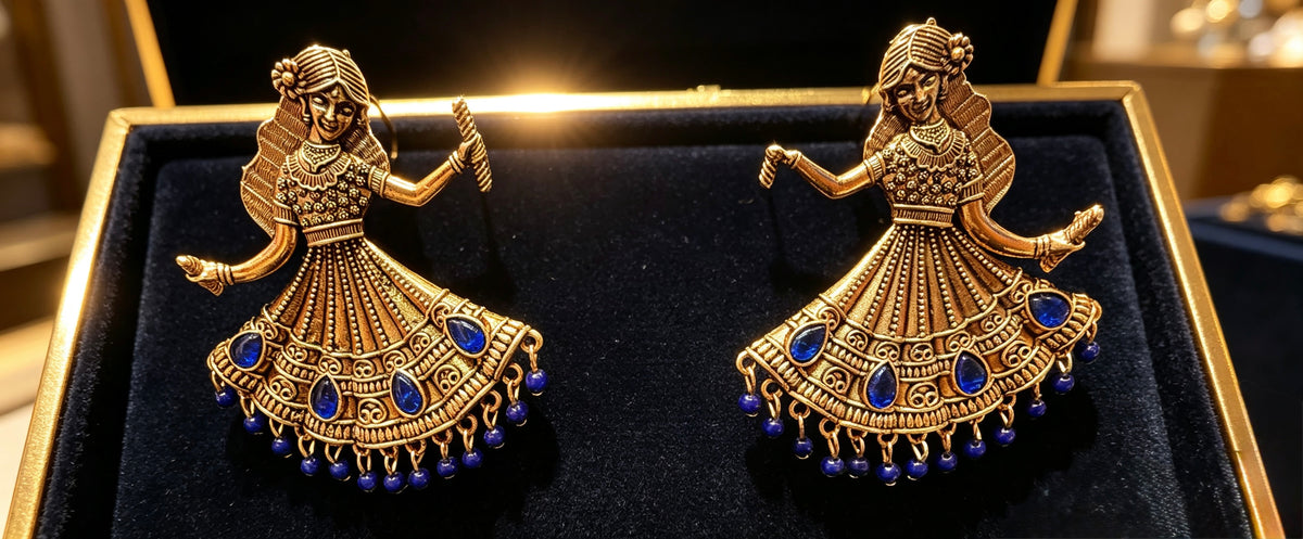 A pair of ornate gold-toned earrings shaped like dancing figures, each decorated with blue stone accents and dangling bead details, displayed on a black velvet jewelry box.