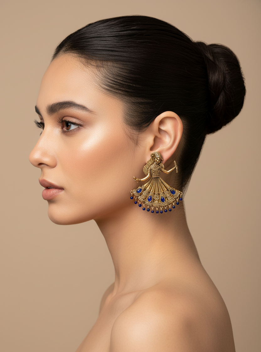 A pair of ornate gold-toned earrings shaped like dancing figures, displayed in a black jewelry box. Each earring features detailed filigree, pink gemstone accents, and dangling pink bead fringe.