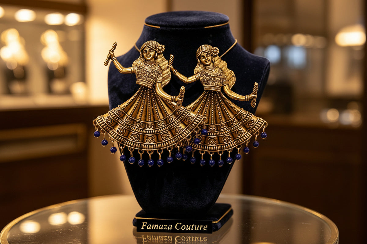 A pair of ornate gold-toned earrings shaped like dancing figures in traditional dress, displayed on a black velvet jewelry bust. The earrings feature intricate detailing, dangling blue bead accents, and are photographed against a softly blurred warm-toned indoor background.
