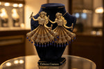 A pair of ornate gold-toned earrings shaped like dancing figures in traditional dress, displayed on a black velvet jewelry bust. The earrings feature intricate detailing, dangling blue bead accents, and are photographed against a softly blurred warm-toned indoor background.