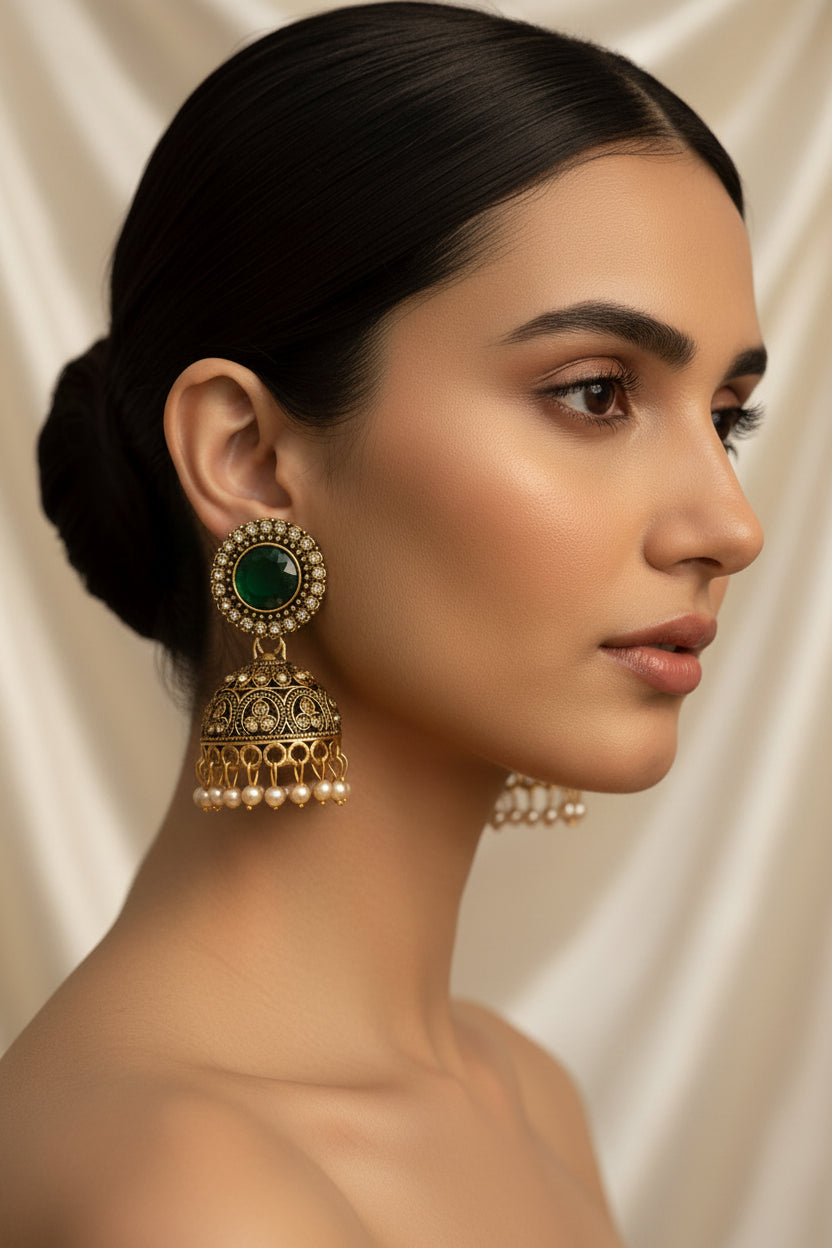 A pair of ornate gold-toned jhumka earrings with green faceted stones and small pearl-like drops, shown hanging from a white jewellery card held by a hand.