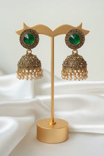 A pair of ornate gold-toned jhumka earrings with green oval stones and rhinestone borders, attached to a white jewellery display card held by a hand.