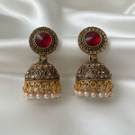 A pair of ornate gold-toned jhumka earrings with red faceted centre stones and small pearl-like drops, displayed on a white jewellery card held by a hand.