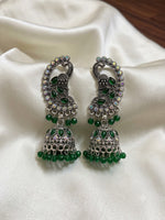 A pair of ornate silver-toned Indian jhumka earrings embellished with green stones and small iridescent rhinestones, displayed on draped white satin fabric.