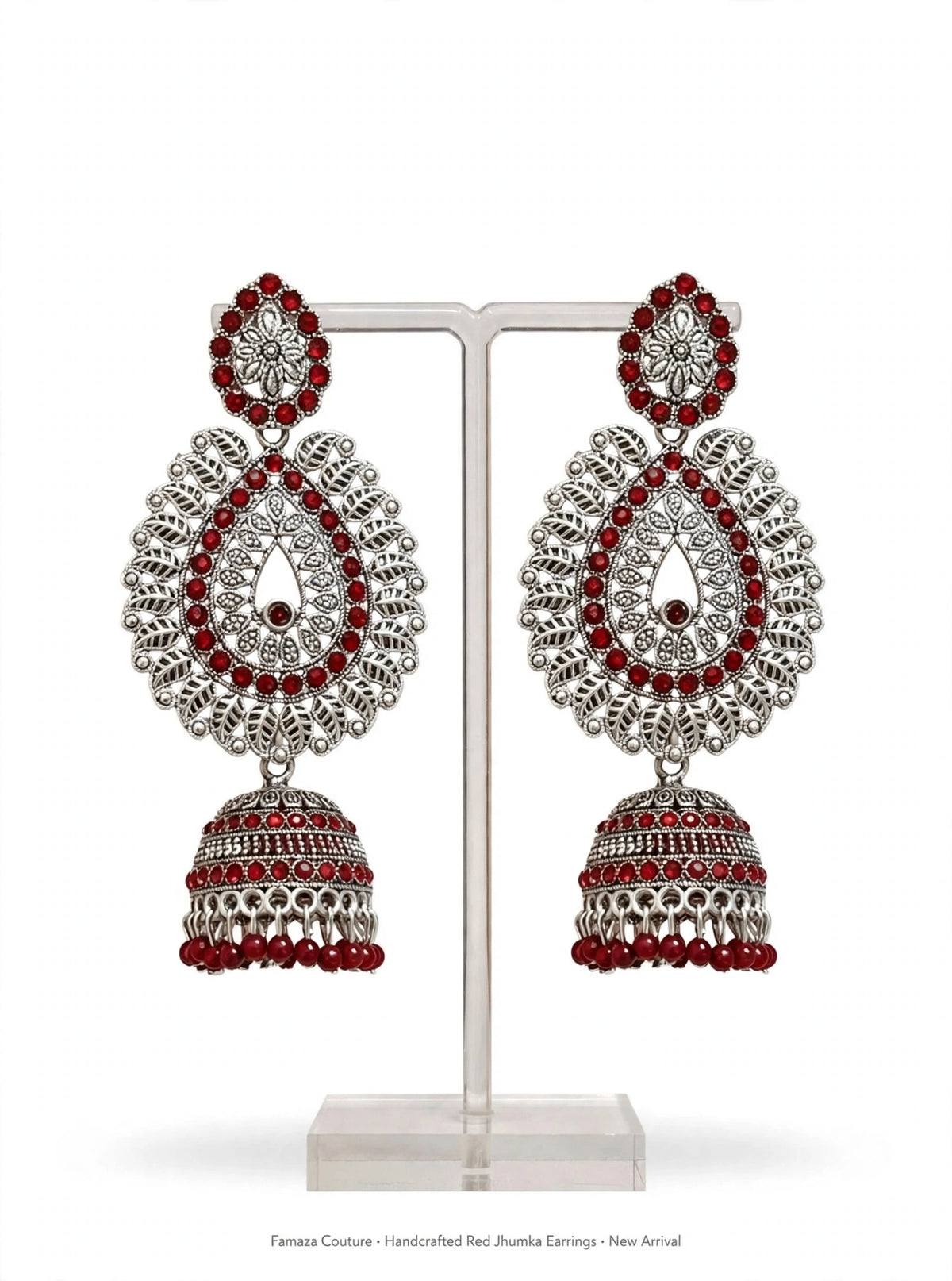 A pair of ornate silver-toned Jhumka earrings with red gemstone accents, displayed on a clear acrylic stand against a white background.