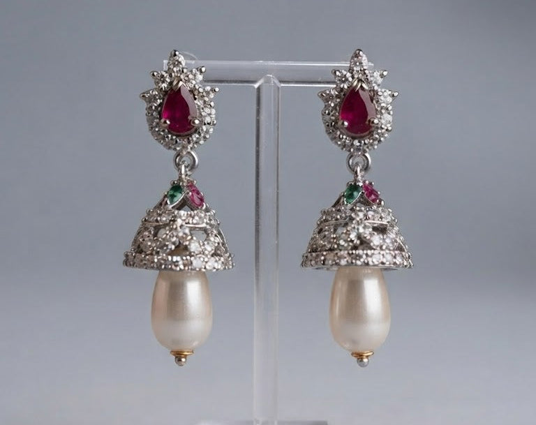 A pair of ornate silver-toned earrings with red teardrop gemstones, small green accent stones, and dangling pearl-like drops, displayed on a clear stand against a soft grey background.