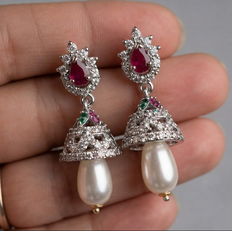 A pair of ornate silver-toned earrings with teardrop-shaped red gemstones, small green and pink accent stones, and dangling pearl-like beads, shown resting on a person's fingers against a soft gray background.