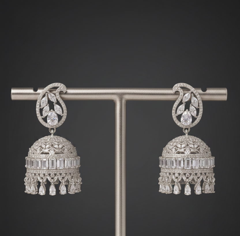 A pair of ornate silver-toned jhumka earrings displayed on a vertical T-stand against a dark gray gradient background. The earrings feature intricate filigree details, a central round stone, and a bell-shaped dangle adorned with rectangular and teardrop-cut stones and hanging tassels.