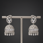A pair of ornate silver-toned jhumka earrings displayed on a vertical T-stand against a dark gray gradient background. The earrings feature intricate filigree details, a central round stone, and a bell-shaped dangle adorned with rectangular and teardrop-cut stones and hanging tassels.