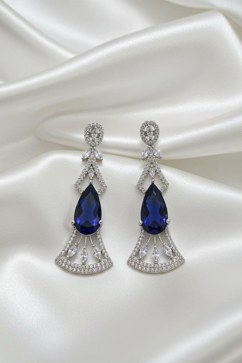 A pair of silver-toned Art Deco style drop earrings with marquise-cut deep blue gemstones and pavé-set clear accent stones, displayed on a black card against a dark background.