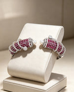 A pair of silver-toned cuff-style earrings with rows of deep red gemstones and small white pearls, displayed on a cream jewelry stand against a warm beige background.