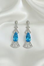 A pair of silver-toned drop earrings with a central faceted blue marquise-cut gemstone, surrounded by smaller clear stones in an Art Deco-inspired fan and leaf design, displayed on a black textured background.
