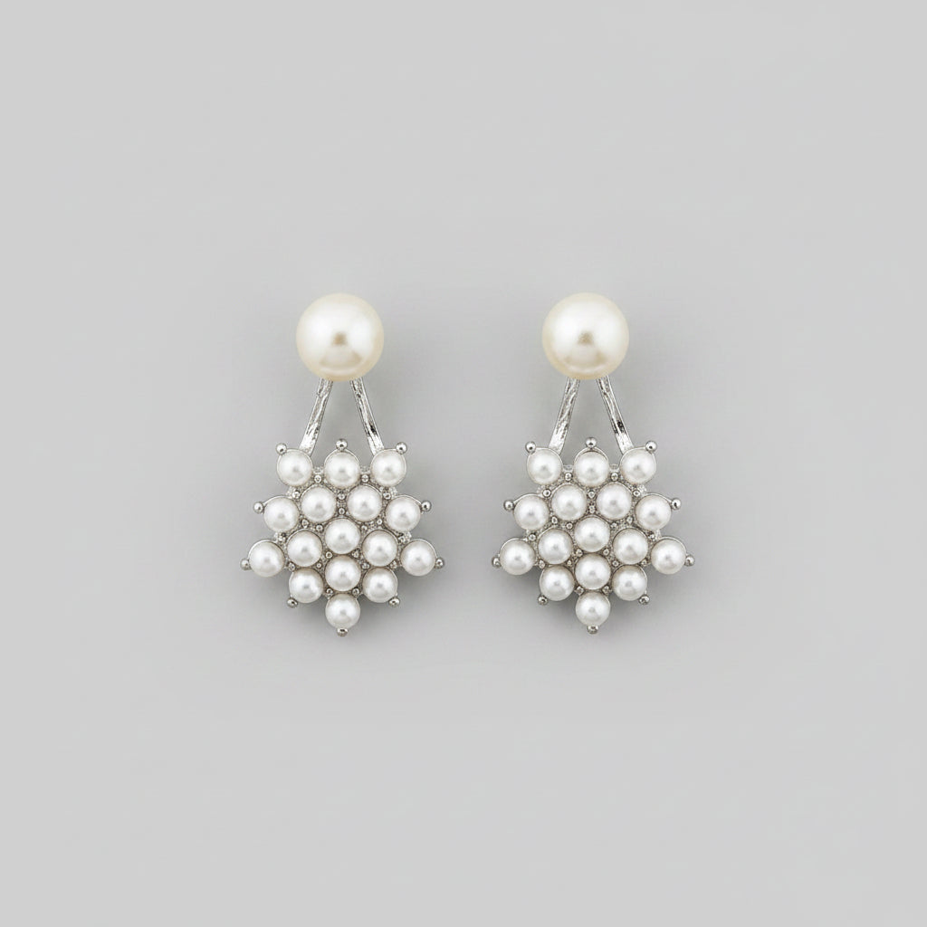 A pair of silver-toned drop earrings with a single round white pearl and a small cluster of tiny silver pearls, displayed on a light wood floor.