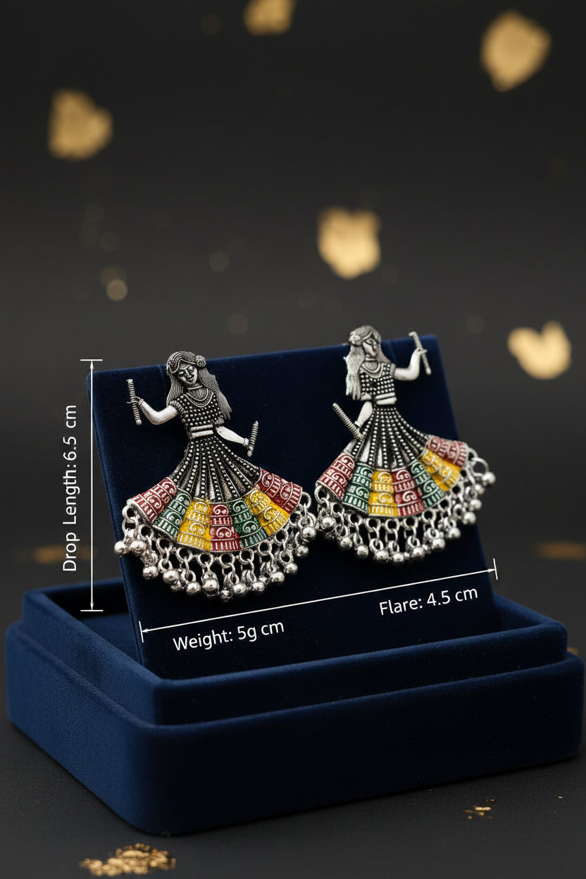 A pair of silver-toned statement earrings shaped like dancing figures with colorful, fan-like skirt details, displayed on a dark blue velvet jewelry stand over a matching box. White measurement labels and text indicate drop length, weight, and flare.