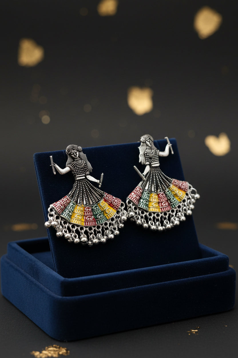 A pair of silver-toned earrings shaped like dancing women in traditional navratri chaniya choli dress, decorated with black beads and colourful enamel details in red, yellow, green, and blue, displayed on a white marble surface.