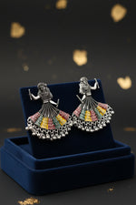 A pair of silver-toned earrings shaped like dancing women in traditional navratri chaniya choli dress, decorated with black beads and colourful enamel details in red, yellow, green, and blue, displayed on a white marble surface.