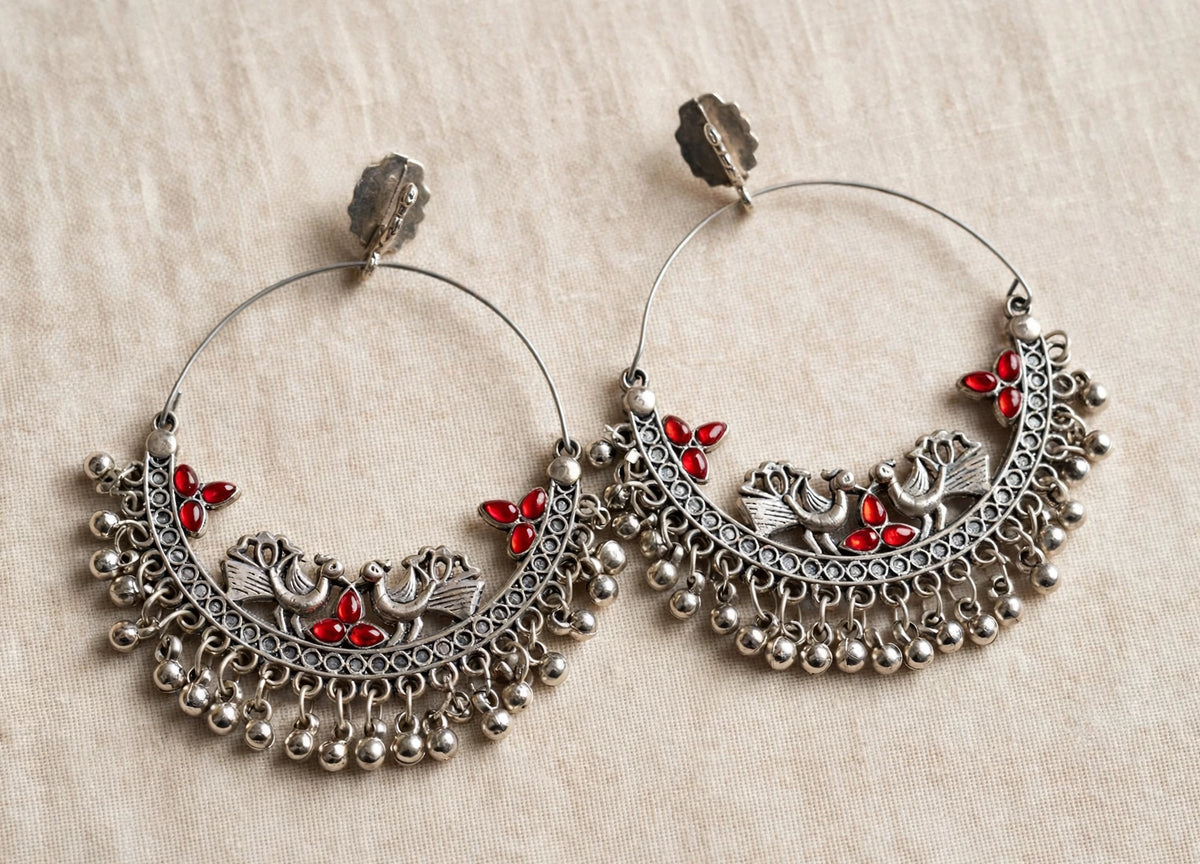 A pair of silver-toned traditional hoop earrings with intricate filigree, bird motifs, and small red stone accents, displayed on a textured beige fabric background.