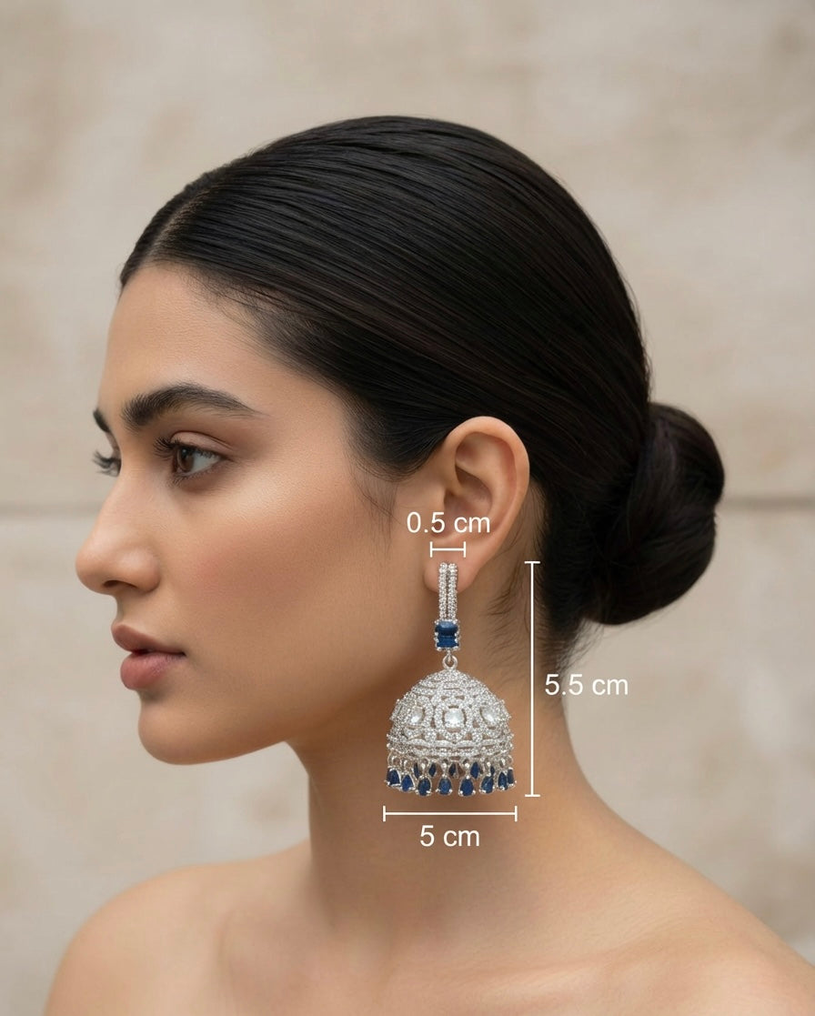 A pair of silver finish Jhumka earrings with American diamonds and blue sapphire stone details, displayed against a plant backdrop