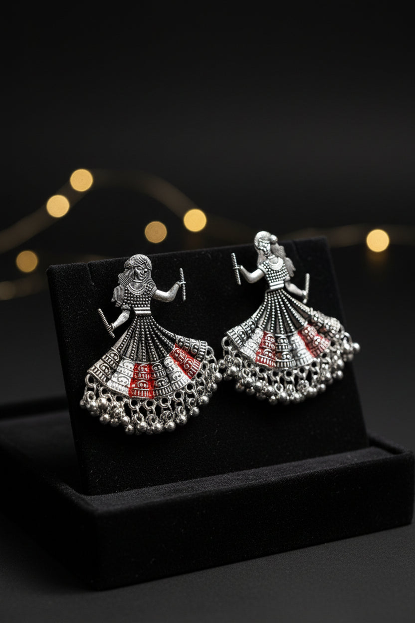 A pair of silver toned metal oxidised earrings design is showing a woman is playing dandiya in navratri lehnga choli traditional dress in red,white and black colour for navratri nights.