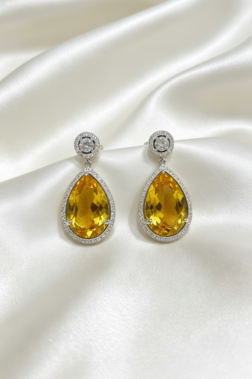 A pair of teardrop-shaped drop earrings with large yellow gemstones in silver settings, displayed on a draped white satin fabric background.
