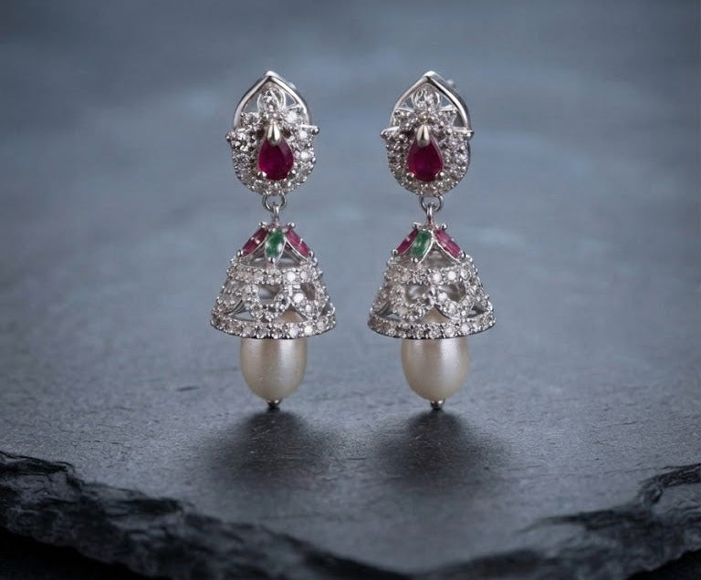 A pair of traditional Indian chandbali earrings with silver-toned metal, red rubies, green emeralds, clear pavé stones, and white pearls, displayed on a dark slate surface.