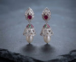 A pair of traditional Indian chandbali earrings with silver-toned metal, red rubies, green emeralds, clear pavé stones, and white pearls, displayed on a dark slate surface.