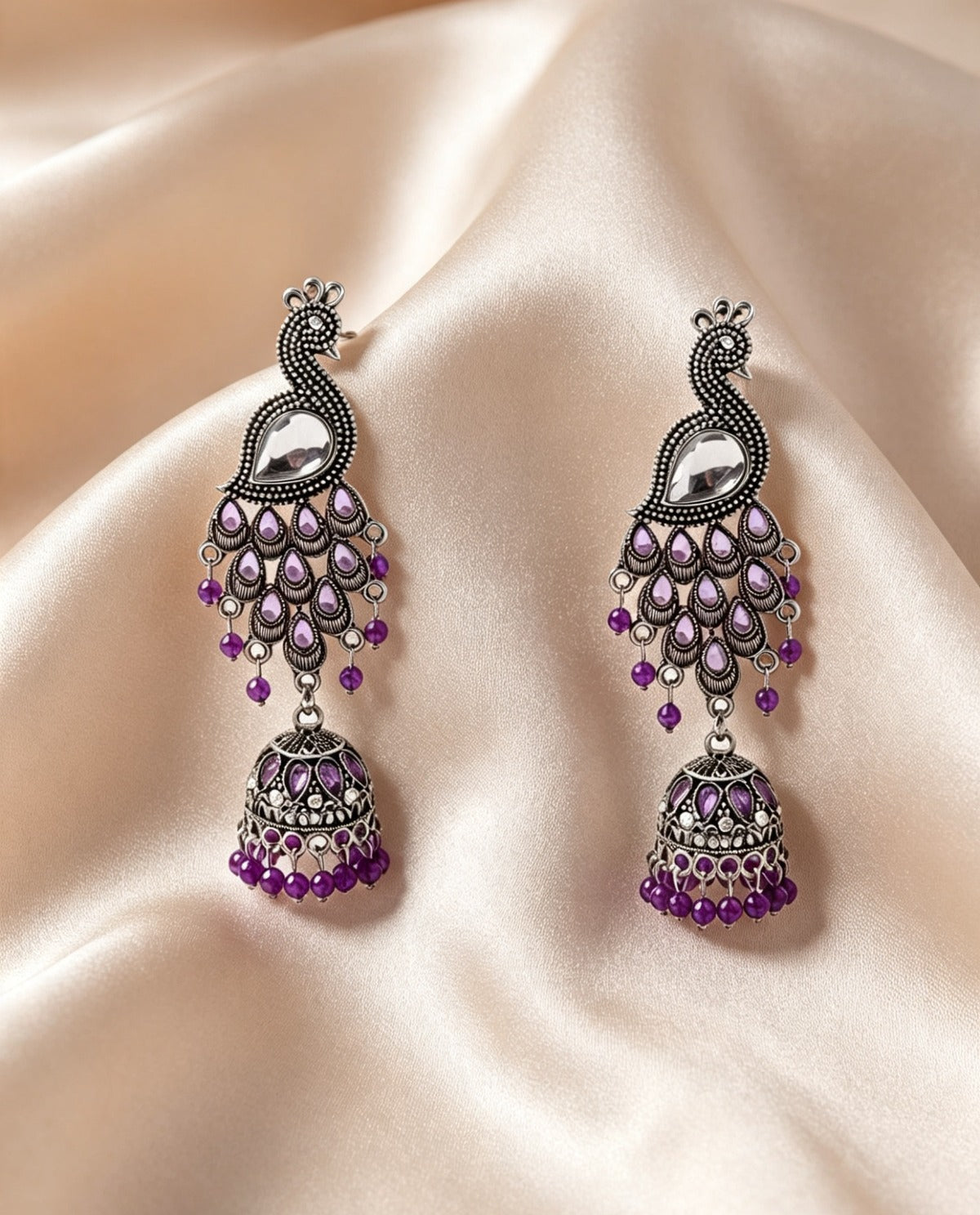 A pair of traditional Indian silver-toned dangle earrings with purple stone embellishments, displayed on a draped beige satin fabric background.