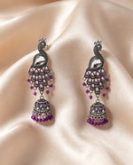 A pair of traditional Indian silver-toned dangle earrings with purple stone embellishments, displayed on a draped beige satin fabric background.