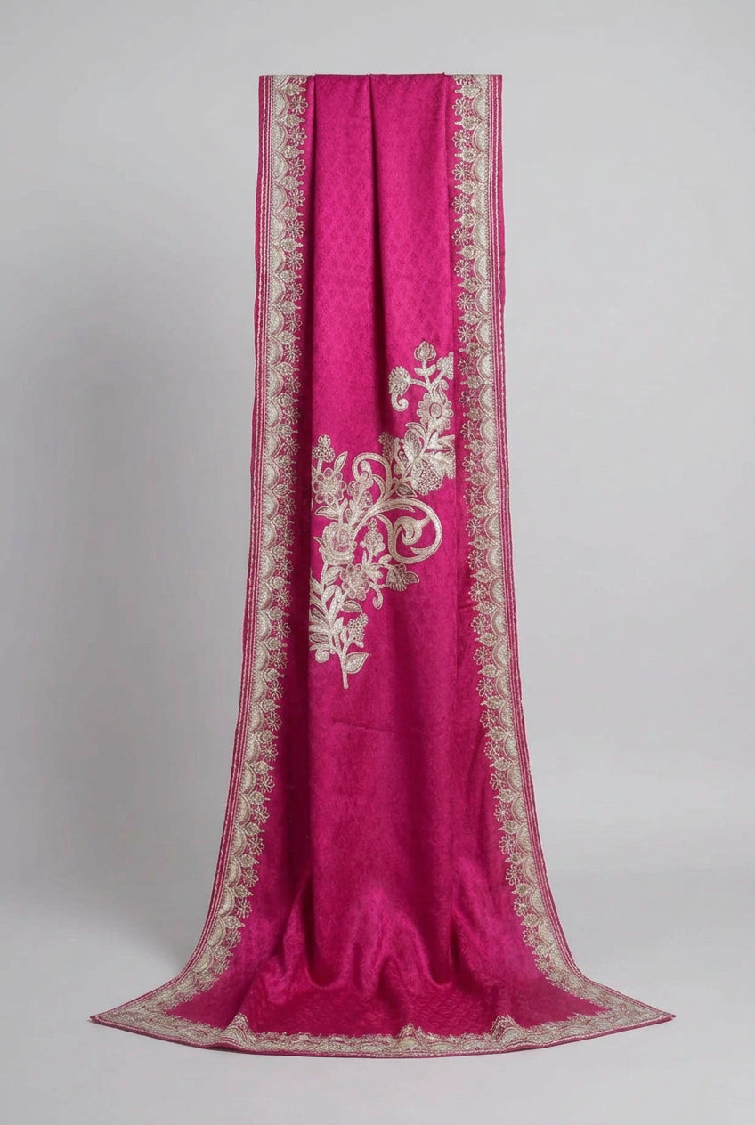A pink Kashmiri Aari embroidered marina scarf with tassels hanging at the ends, displayed on a wooden hanger with greenery in the background.