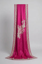 A pink Kashmiri Aari embroidered marina scarf with tassels hanging at the ends, displayed on a wooden hanger with greenery in the background.