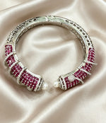 A silver-toned cuff bracelet featuring rows of deep pinkish-red round gemstones and small white pearl accents, displayed on draped champagne satin fabric.