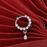 A silver-toned huggie hoop earring embellished with clear round stones and a dangling teardrop-shaped clear stone, displayed on a red velvet surface.