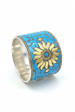 A silver-toned mosaic cuff bracelet featuring a central gold-toned sunburst design over a background of bright turquoise blue mosaic tiles, set against a dark textured surface.