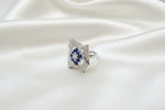 A silver-toned ring with a rectangular pavé setting and small blue gemstones, photographed on a textured blue fabric background.