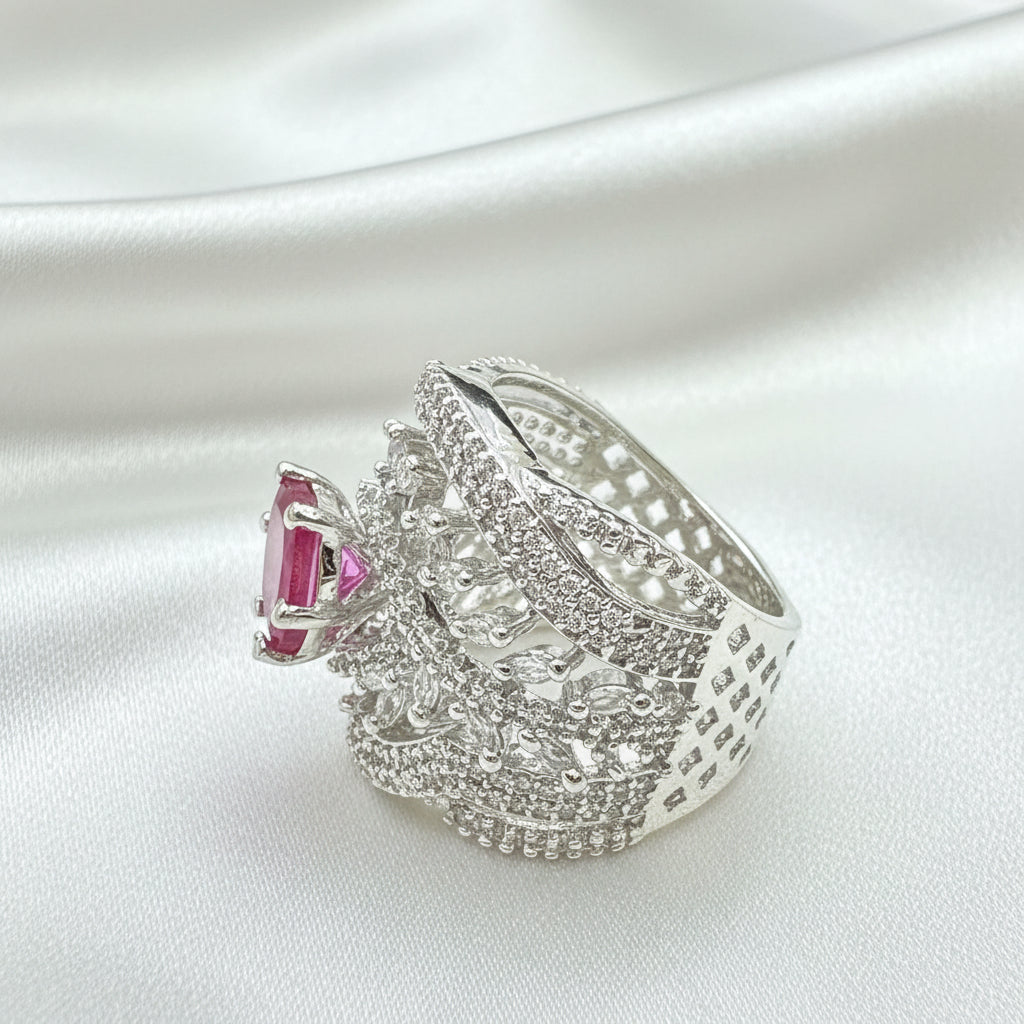 A silver-toned statement ring featuring a large, ornate pavé-set design with a prominent rectangular pink gemstone on the side, resting on a dark textured surface.