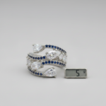 A silver-toned wrap bracelet with pear-cut clear stones and rows of deep blue round stones, shown on a light gray background next to a small digital scale displaying 5 g.