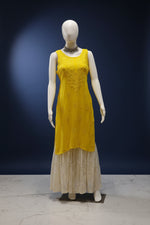 A sleeveless bright yellow kurta dress displayed on a white mannequin, paired with a cream-colored embroidered skirt. The mannequin is positioned in front of a dark glass door decorated with red and white flower garlands.