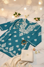 A teal blue tunic top with white floral and leaf block prints, hanging on a wooden hanger against a white draped fabric background.