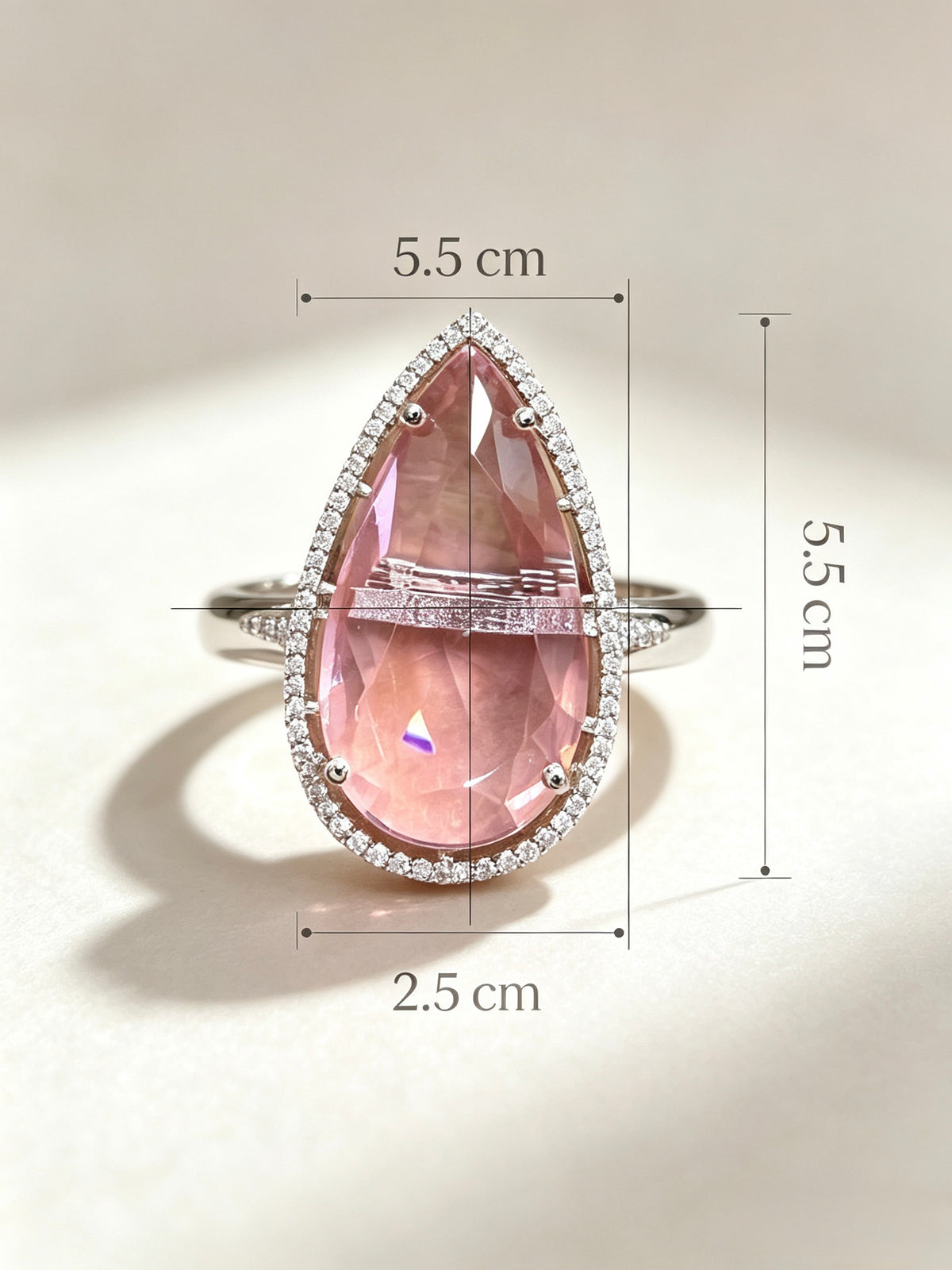 A teardrop-shaped pink gemstone ring with a silver-toned halo and band, shown on a light beige background with overlaid dimension lines and text indicating 5.5 cm width, 5.