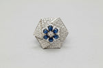 A vintage-style hexagonal brooch featuring a silver-toned pavé setting with a central blue floral motif, resting on a textured blue velvet surface.