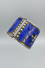 A wide cuff bracelet worn on a wrist, featuring a mosaic of vivid blue stone pieces with gold-tone studs and silver-tone beaded trim.