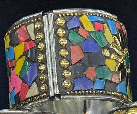 A wide silver-toned cuff bracelet featuring a colorful mosaic inlay of geometric shapes in red, blue, green, yellow, pink, and black, accented with rows of gold-tone studs and a gold-tone floral motif on the right side.
