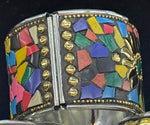 A wide silver-toned cuff bracelet featuring a colorful mosaic inlay of geometric shapes in red, blue, green, yellow, pink, and black, accented with rows of gold-tone studs and a gold-tone floral motif on the right side.