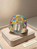 A wide silver cuff bracelet with a colourful mosaic floral design, displayed on a beige velvet jewellery box. The bracelet features a silver-toned band, a beaded edge, and a top half decorated with a multicolour mosaic pattern of yellow, blue, red, green, and pink tiles surrounding a gold-toned flower motif.