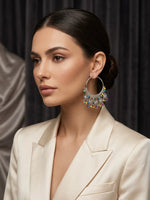 A woman in a cream satin blazer wearing a large silver-toned hoop earring adorned with colourful rhinestones and dangling crystal elements, set against a dark curtain background.