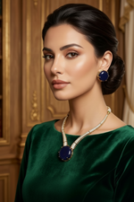 A woman with dark hair styled in a low bun wears a luxurious gold necklace and matching gold earrings with deep blue oval stones, paired with a dark green velvet top against a warm, ornate gold-paneled background.