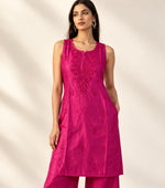 A woman modelling a bright pink kurta set with matching trousers, featuring embroidered detailing on the chest, set against a textured white curtain backdrop.