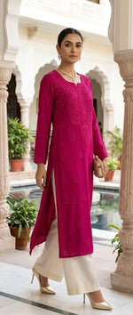 A woman models a magenta pink embroidered kurta with cream wide-leg pants in an outdoor courtyard setting with white pillars and a pool in the background.