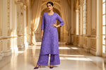 A woman models a purple embroidered kurta set with matching wide-leg pants in an ornate, sunlit palace corridor with beige drapes and gold architectural details.