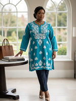 A woman models a teal blue kurta with white floral embroidery, paired with navy blue trousers and beige heels, standing in a bright room with large arched windows.